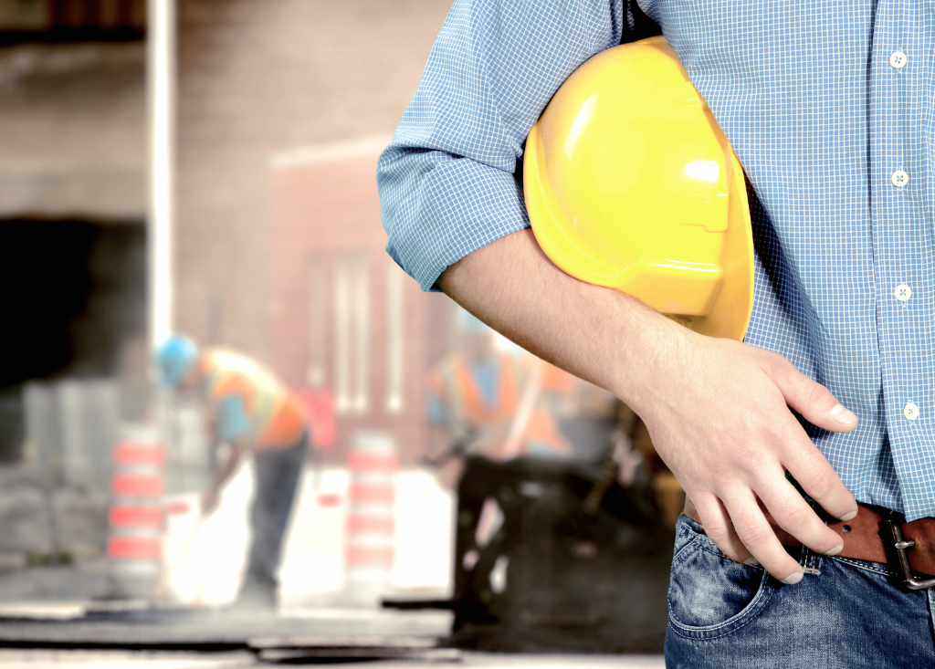 Mężczyzna w koszuli trzymający kask ochronny na tle pracowników budowy – symbolizujące prawo pracy i sprawy pracownicze.